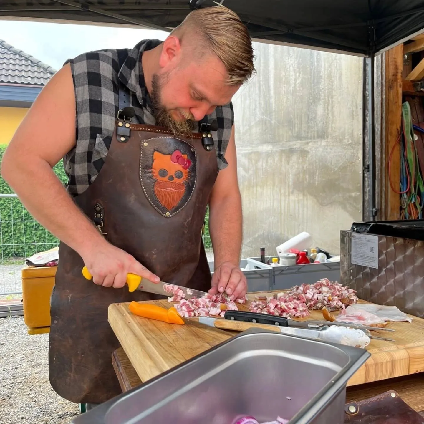 Raphael Fössl in Lederschürze schneidet auf einem Holzbrett Speck für den Grillkurs